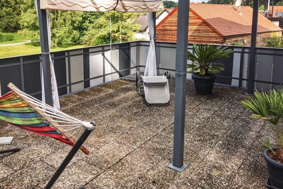 La terrasse de toit avec dalles en béton ancien offre un espace de séjour généreux. La surface sale des dalles montre des signes d'usure visibles. Le store offre une ombre agréable lors des journées chaudes. Les pots de plantes avec des palmiers et un hamac créent une ambiance accueillante. La structure de garde-corps en métal et verre complète le design moderne et ouvert de la terrasse.