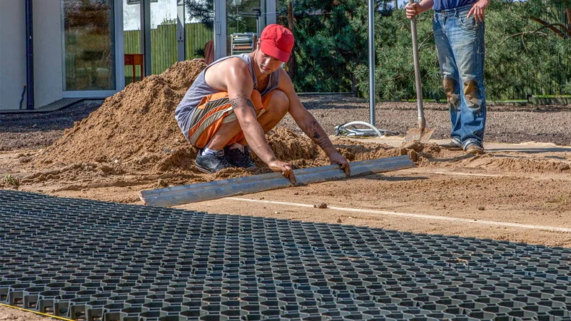 Nel giardino, uno strato di base di griglie di plastica a nido d'ape viene posato sul letto di sabbia. Un uomo livella la sabbia.