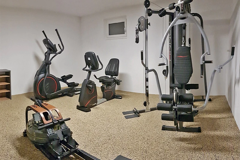 Das Bild zeigt ein Homegym, das mit einem hochwertigen WARCO-Fitnessboden im Farbdesign Travertin ausgestattet ist. Der Bodenbelag besteht aus einem robusten Material, das für Langlebigkeit und hohe Belastbarkeit sorgt und gleichzeitig eine ansprechende Optik bietet. Der Raum ist mit verschiedenen Fitnessgeräten ausgestattet, darunter ein Rudergerät, ein Crosstrainer, ein Ergometer und eine Kraftstation. Die helle, natürliche Farbgebung des Travertin-Designs harmoniert gut mit den modernen Fitnessgeräten und verleiht dem Raum eine warme und einladende Atmosphäre. Die Wände des Raumes sind in schlichtem Weiß gehalten, was den Fokus auf die Funktionalität und den Komfort des Fitnessbereiches lenkt.