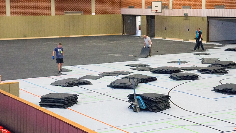 In un grande palazzetto dello sport viene posato il pavimento per eventi WARCO. Si tratta di tappeti in granulato di gomma con un design di pavimentazione ad incastro. Il pavimento per eventi protegge il delicato pavimento sportivo della sala da eventuali danni.