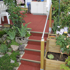 rote Treppe führt auf Terrasse. Rund um den WARCO-Treppenbelag wachsen grüne Pflanzen.