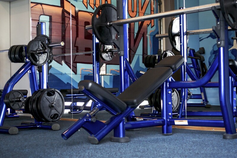 In einem Fitnessstudio blickt man auf eine blau lackierte Schrägbank, die vor einer Spiegelwand steht. Die Schrägbank steht auf einem blauen Bodenbelag. Dabei handelt es sich im Fitness Bodenschutzmatten von WARCO im Farbdesign Atlantik.
