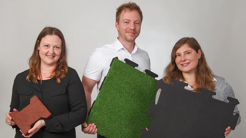 Lächelnd präsentieren drei Fachberater von WARCO verschiedene Bodenplatten aus pu-gebundenem Gummigranulat in Braun, Grün und Schwarz. Diese Platten eignen sich besonders für Balkone und bieten rutschhemmende, langlebige Eigenschaften. Jede Oberfläche hat ein eigenes Design und lässt sich einfach verlegen. Mit ihrer kompetenten Beratung unterstützen die Experten bei der Wahl des passenden Bodenbelags, damit jeder Balkon sicher und ansprechend gestaltet werden kann. Die pflegeleichten Platten überzeugen durch Strapazierfähigkeit und Komfort.