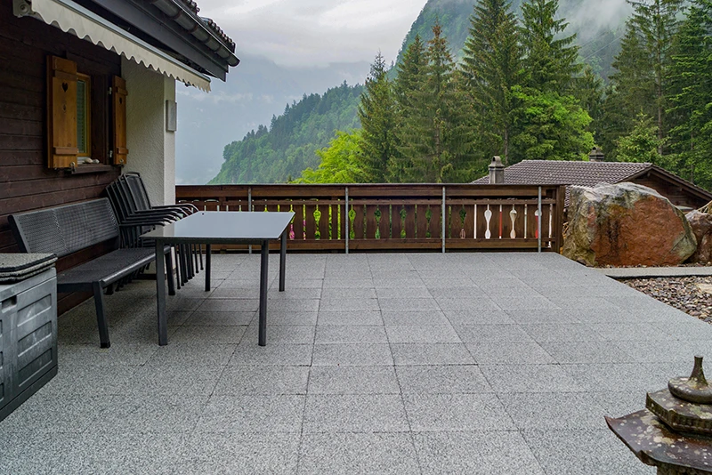 Piastrelle quadrate WARCO in gomma con finitura grigio marmorizzata costituiscono la pavimentazione di una terrazza con le montagne sullo sfondo. Le piastrelle sono posate con schema regolare e accostate con giunti ridotti. La terrazza è direttamente a ridosso di un edificio con facciata in legno.