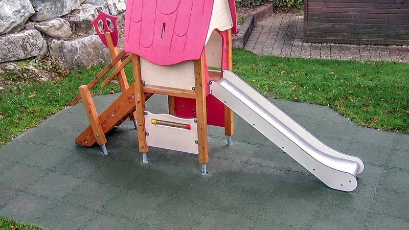 Auf einem Hanggrundstück wurde vor der Stützmauer ein Kleinkinderspielplatz, bestehend aus einem Kleinkinderspielhaus mit Rutsche, errichtet. Der Fallschutz erfolgt fachgerecht mit Fallschutzmatten nach EN 1177.