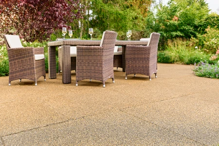 Terrazza soleggiata in giardino con piastrelle WARCO beige-marrone in granuli di gomma legati in PU. La superficie antiscivolo e permeabile all’acqua garantisce un’eccellente comodità di camminata e riduce i rumori. Un robusto tavolo da giardino con sedie intrecciate e calici di vino invita al relax. Arbusti rigogliosi e fiori variopinti creano un ambiente naturale. Questo pavimento elastico resiste al gelo, mantiene la forma e si adatta all’uso in ogni stagione.