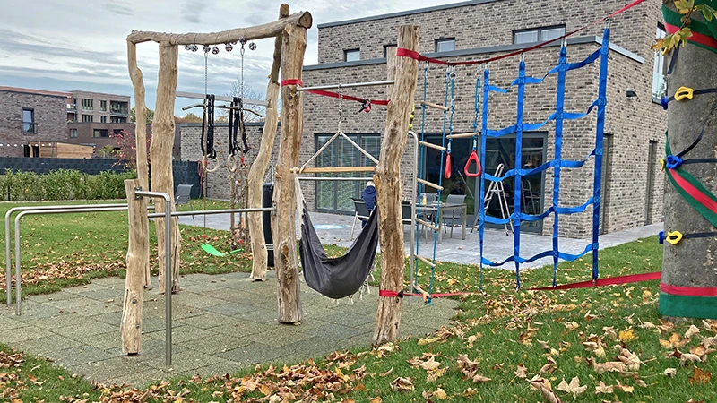 La struttura per arrampicarsi è attaccata a un albero con delle reti. Accanto, alcuni pali di legno con un sacco da boxe, una sbarra alta e un'altalena.