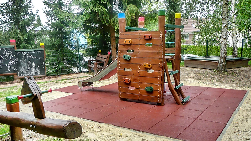 Fallschutz aus roten Fallschutzplatten rund um Kletterturm auf Spielplatz, wartungsfrei und stoßdämpfend.