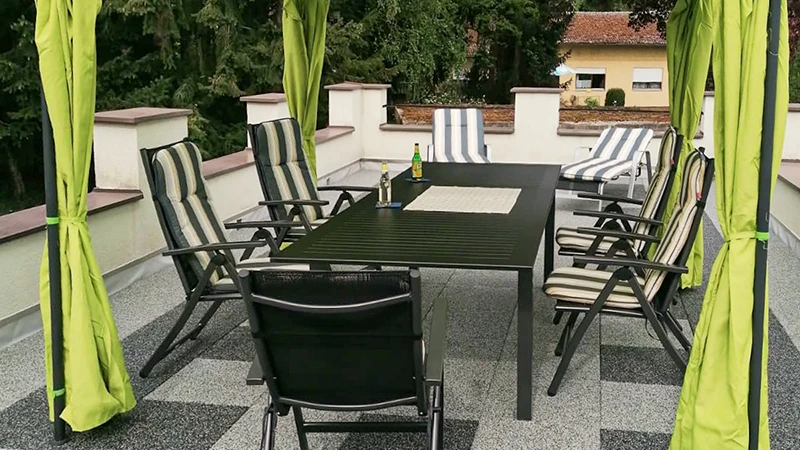 Su un terrazzo sul tetto è posata una pavimentazione realizzata con piastrelle da terrazza WARCO, caratterizzate da una venatura grigio chiaro e scuro che ricorda il granito. Un baldacchino copre l’area salotto sulla superficie elastica e resistente alle intemperie. Le piastrelle antiscivolo e ammortizzanti si installano senza adesione permanente e attenuano i rumori da calpestio. Il risultato è uno spazio di facile manutenzione, ideale per momenti di relax all’aperto.