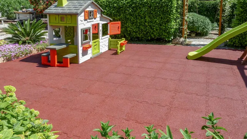 Une aire de jeux pour les enfants dans le jardin. Sur un vaste terrain en dalles amortissantes rouges de sécurité WARCO a été installée une maisonette en plastique.