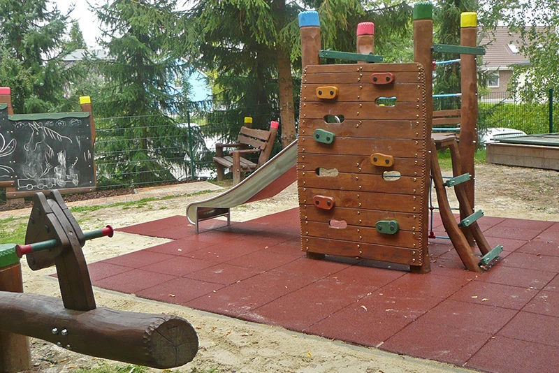 Das Bild zeigt einen naturnah gestalteten Spielplatz, der mit ziegelroten WARCO Fallschutzmatten ausgestattet ist. In der Bildmitte befindet sich ein Spielgerät aus Holz, bestehend aus einer Kletterwand mit bunten Griffen, einer Rutsche und weiteren Kletterelementen. Der Bereich um das Klettergerüst ist mit Fallschutzmatten ausgelegt, die eine sichere und stoßdämpfende Oberfläche bieten. Die Umgebung des Spielplatzes ist von hohen Nadelbäumen und Sträuchern umgeben, die eine naturnahe Atmosphäre schaffen. Im Hintergrund ist eine Tafel zu sehen, auf der Kinder mit Kreide gemalt haben. Der Spielplatz bietet eine harmonische Kombination aus natürlicher Gestaltung und modernen Sicherheitsmaßnahmen.
