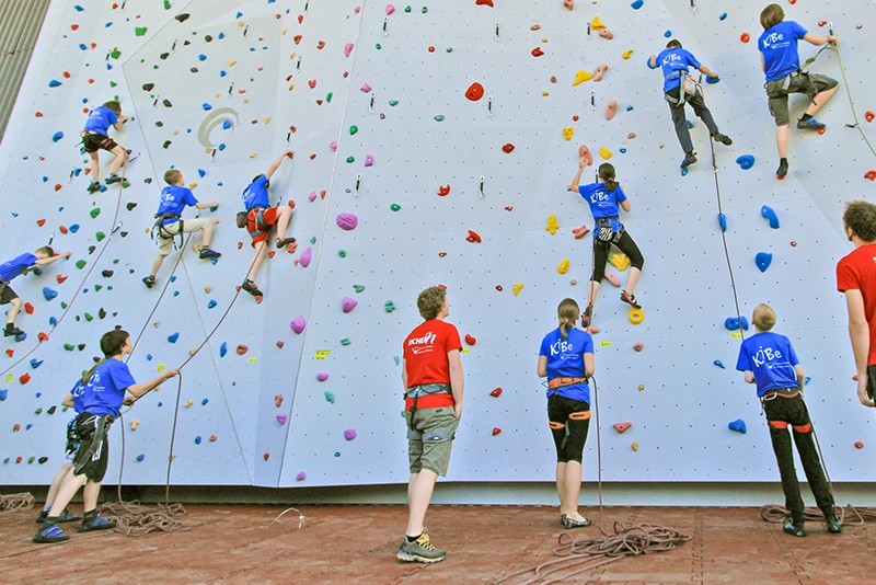 Des enfants et adolescents pratiquent l’escalade dans une salle intérieure équipée d’un mur d’escalade avec cordes de sécurité. Le sol est recouvert de dalles en caoutchouc antichoc rouges de WARCO, qui offrent une surface amortissante pour réduire les risques de blessure en cas de chute. Ces dalles emboîtables assurent une installation stable et durable, idéale pour les salles de sport et les centres d’escalade.