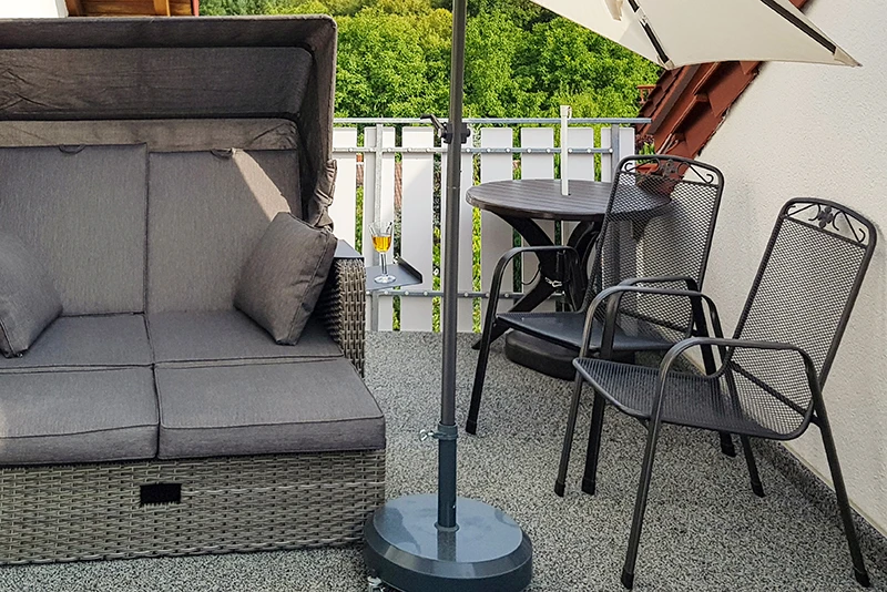 Auf dem Foto sieht man einen Balkon, der mit grauen WARCO-Bodenplatten ausgelegt ist. Ein bequemer Strandkorb sowie ein kleiner Tisch mit zwei Metallstühlen bieten Platz zum Entspannen und Genießen. Der elastische und rutschfeste Bodenbelag schützt vor Feuchtigkeit und sorgt für Trittschalldämmung. Gleichzeitig ist er pflegeleicht und witterungsbeständig, sodass der Außenbereich lange schön bleibt und zum Verweilen einlädt.