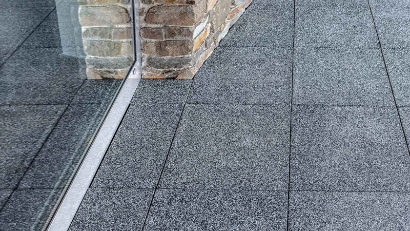 Auf einer Terrasse liegen quadratische Terrassenplatten im Farbdesign Grauer Granit. Die Terrassentür grenzt an eine schräg verlaufende Natursteinmauer aus Sandstein. Die Terrassenplatten von WARCO sind passend zum Profil der Wand zugeschnitten und liegen bündig ohne Fuge an.