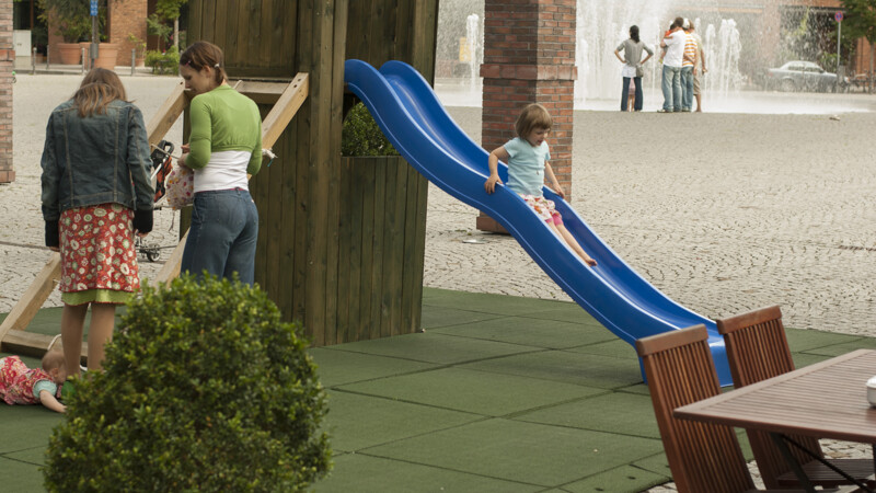Auf dem gepflasterten Platz hat der Gastwirt einen Spielplatz für die Kinder seiner Gäste im Gartenlokal angelegt. Der Spielplatz, auf dem sich ein Kletterturm aus Holz mit Rutsche befindet, ist mit dem mobilen WARCO-Spielboden gesichert.