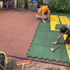 Ein Mann verlegt grüne Fallschutzplatten mit Puzzleverzahnung, d.h. in die Seiten eingeformtem Schwalbenschwanz-Verbindungssystem. Die Fallschutzplatten werden auf einer Tragschicht aus Kunststoffwabengitter verlegt. Die planeben verlegten Wabengitter sind mit rotem Splitt gefüllt.