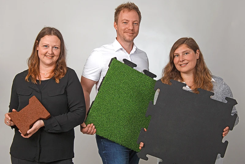 Drei WARCO-Mitarbeiter präsentieren verschiedene Fallschutzplatten. Von links nach rechts: Frau Velic hält ein Verbundpflaster-Element, das für befahrbare Flächen verwendet wird. Herr Sinner zeigt eine Fallschutzplatte mit Kunstrasenbelag, ideal für Indoor-Spielplätze. Frau Sperling präsentiert eine schwarze Puzzleplatte, die leicht zu verlegen ist und in verschiedenen Varianten hergestellt wird. Diese Produkte zeigen die Vielfalt und Qualität der WARCO Fallschutzlösungen.