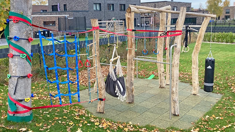 La struttura di arrampicata per bambini con le lastre antitrauma WARCO è attaccata a un albero con delle reti. Accanto, alcuni pali di legno con un sacco da boxe, una sbarra alta e un’altalena.