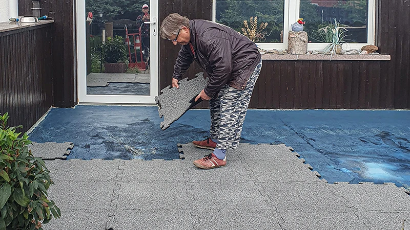 Eine Terrasse wird renoviert. Eine ältere Dame verlegt die nächste 18 mm dicke Terrassenplatte mit Puzzleverzahnung auf die bereits teilweise mit neuen Platten belegte Terrasse. Eine Abdichtungsfolie dient als Untergrund für den neuen Bodenbelag aus Gummiplatten. Die passgenaue Verzahnung der Platten ermöglicht eine einfache und stabile Verlegung.