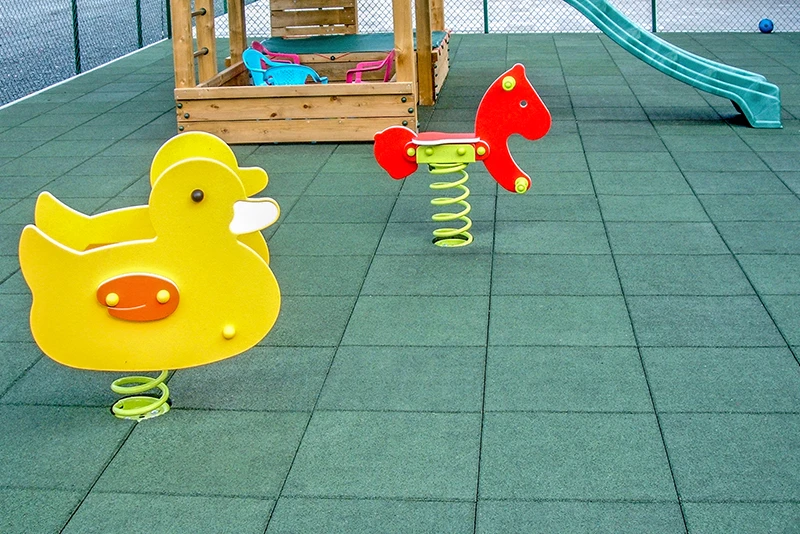 Das Bild zeigt einen Spielplatz, dessen Boden mit grasgrünen WARCO Fallschutz-Gummimatten ausgelegt ist. Im Vordergrund sind zwei bunte Wipptiere auf Federn zu sehen: eine gelbe Ente und ein rotes Pferd. Im Hintergrund befindet sich eine kleine Holzstruktur mit bunten Sitzgelegenheiten und einer grünen Rutsche. Der gesamte Spielplatz ist von einem Maschendrahtzaun umgeben, der für Sicherheit und Schutz sorgt. Die Fallschutzmatten bieten eine stoßdämpfende Oberfläche, die die Kinder vor Verletzungen schützt und gleichzeitig für ein attraktives grünes Erscheinungsbild des Spielplatzes sorgt.
