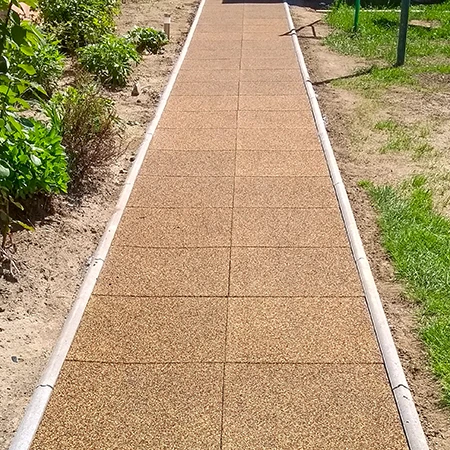 Un vialetto da giardino dritto in piastrelle quadrate di gomma con cordolo laterale attraversa l’aiuola. Le piastrelle marroni in granuli di gomma offrono una superficie antiscivolo, resistente alle intemperie e facile da mantenere. Il vialetto garantisce un passaggio sicuro e pulito in ogni condizione. Ideale per giardini, cortili e accessi.