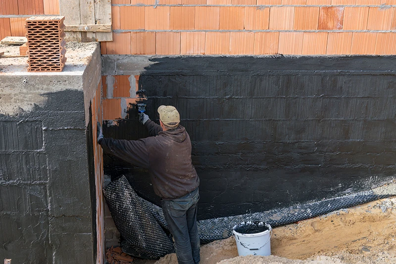 Ein Handwerker trägt ALLESDICHT von WARCO, eine elastische Gummiabdichtung, auf die Außenwand eines neu errichteten Weinkellers auf. Die wasserverdünnbare Abdichtung wird mit einem Pinsel aufgetragen und bildet nach dem Trocknen eine absolut wasserdichte und reißfeste Gummihaut. Im Bild sind unverputzte Ziegelwände sowie die Abdichtungsmasse in einem Eimer am Boden zu sehen. Die Abdichtungsarbeiten finden statt, bevor die Baugrube verfüllt wird, um das Bauwerk vor eindringendem Wasser zu schützen.