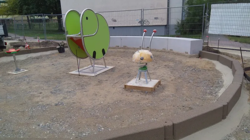 Ein Kleinkinderspielplatz in Form einer runden Insel befindet sich im Bau. Die Spielgeräte sind bereits aufgestellt, die Tragschicht für den Boden ist eingebaut. Die Fläche wird mit Kettenelementen, d.h. miteinander verbundenen Blockstufen aus Gummigranulat, eingefasst.
