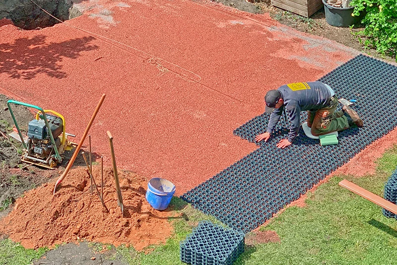 Un uomo sta posando i pavimenti sportivi tipo puzzle di WARCO di colore verde erba sulle griglie a nido d’ape in plastica riempite di ghiaia.